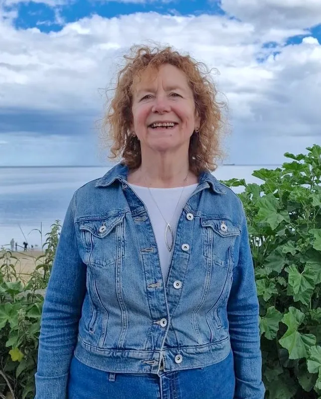 Diane Boote smiling outdoors in a denim jacket, with the sea and a cloudy sky behind her.
