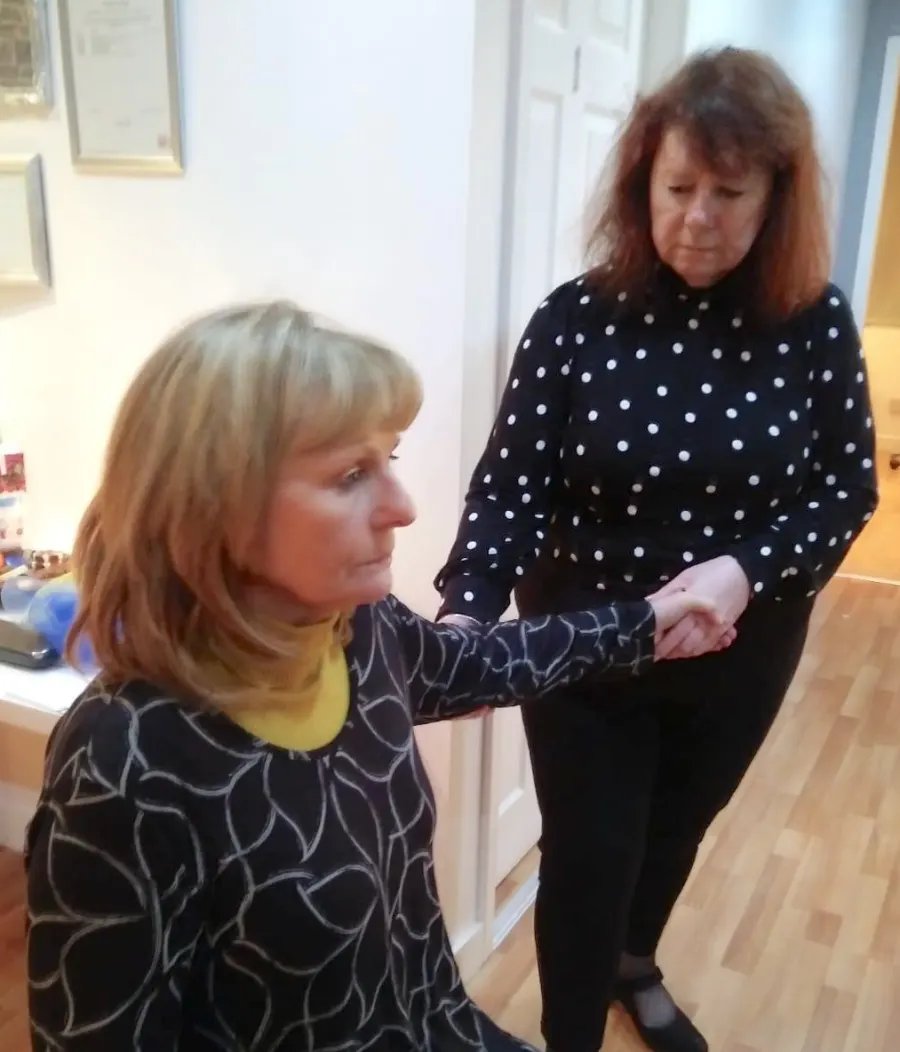 Diane, standing, gently holds the forearm and hand of a seated woman, who looks ahead calmly.